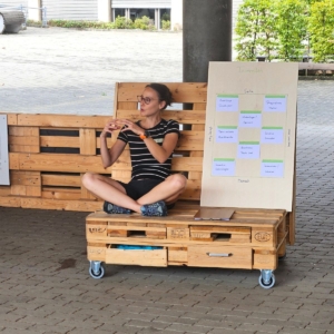 Melanie Wabro sitzend auf einem Palettenmöbel hält einen Entwicklungsimpuls zu Fähigkeiten & Stärken im Team mit einer Holztafel.
