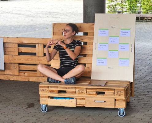 Melanie Wabro sitzend auf einem Palettenmöbel hält einen Entwicklungsimpuls zu Fähigkeiten & Stärken im Team mit einer Holztafel.