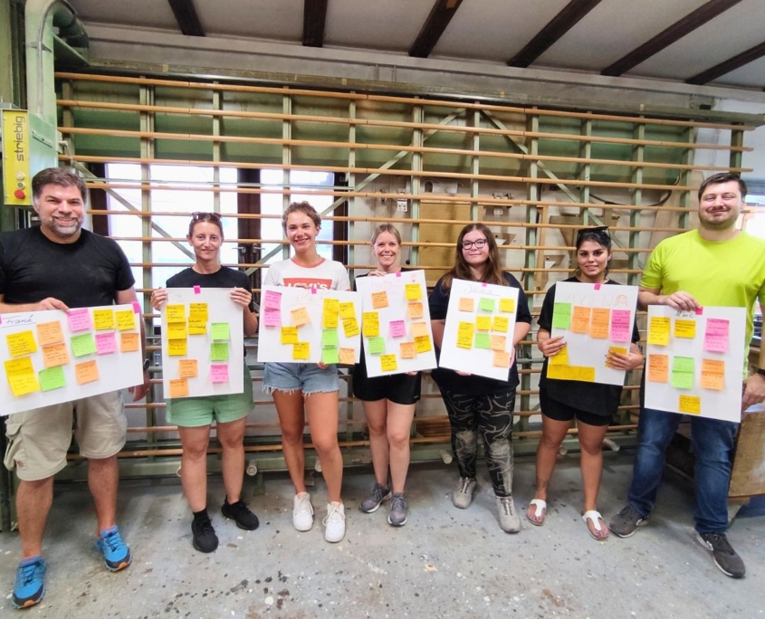 Abschluss Teambuilding-Workshop. Team steht vor der Plattensäge ind er Werkstatt mit Plakaten. Reflexion der Teamrollen von cbs Corporate Business Solutions